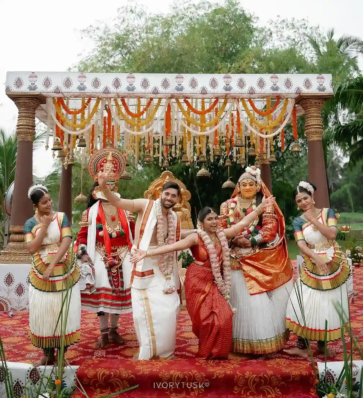 Bride and groom posing with Kathakali artists and Mohiniyattam dancers on a decorated outdoor mandap stage during a destination Kerala wedding.