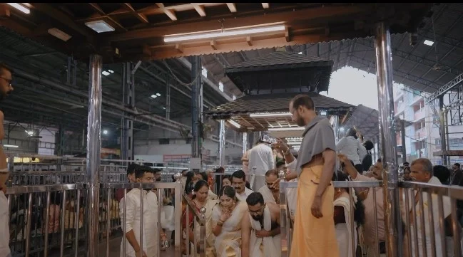 Bride in Kasavu saree and groom in traditional Kerala attire entering the covered Guruvayur Temple kalyana mandapam stage area surrounded by family and Devaswom staff.
