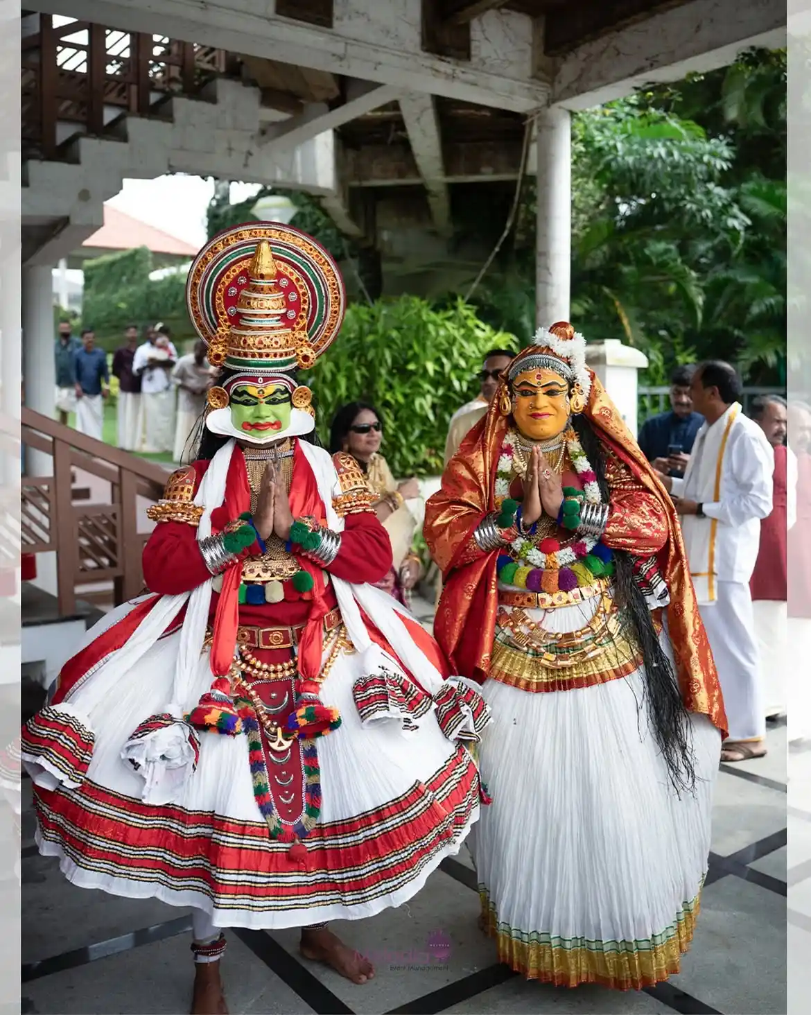 Two Kathakali artists in full traditional costume and facial makeup standing together in a Namaste pose at Le Méridien Kochi.