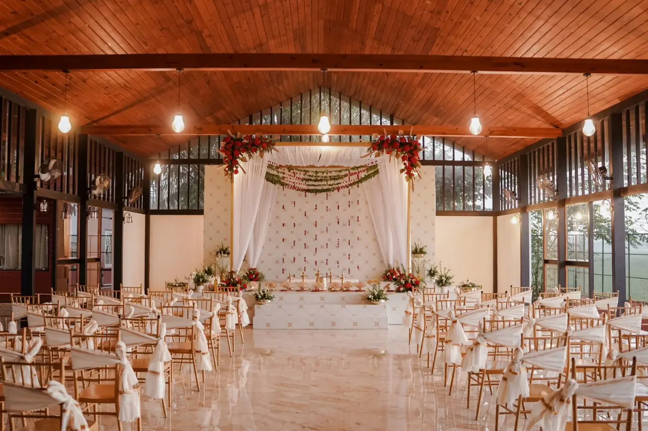 Aesthetic indoor wedding stage decoration by Melodia Event Management featuring elegant white drapes, red floral arrangements, warm hanging bulbs, and wooden Chiavari chairs under a vaulted ceiling.
