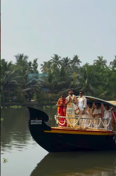 Bride and groom in traditional Kerala attire riding a decorated wooden house boat on calm backwaters surrounded by coconut trees.