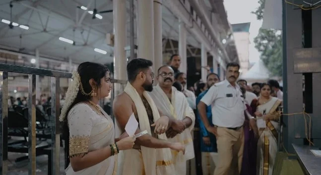 A bride and groom in traditional Kerala wedding attire holding their advance booking receipt while waiting in line at the submission office near Melppathur Auditorium in Guruvayur Temple.