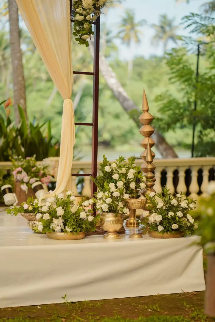 Outdoor riverside wedding stage setup featuring white rose floral arrangements in golden Urli bowls and beige fabric drapes at Kanjiras Luxeves Pavilion in Triprayar, Thrissur.