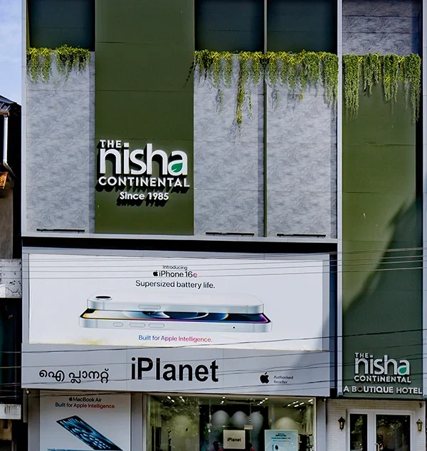 Exterior facade of The Nisha Continental boutique hotel featuring modern architectural panels, an iPlanet Apple reseller store on the ground floor with an iPhone billboard above it, and lush green hanging plants along the upper exterior wall.