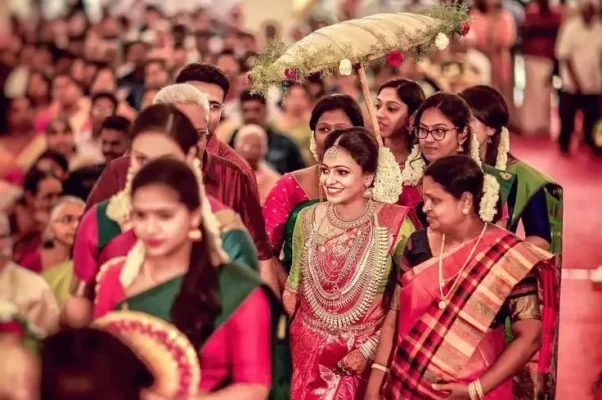 A vibrant indoor traditional Kerala wedding ceremony at Guruvayur with around 500 seated guests watching the bride and groom exchange garlands on a floral-decorated stage.