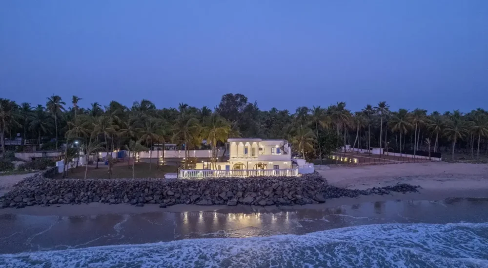 An aerial twilight view of a coastal property featuring a whitewashed, Mediterranean-style multi-story villa lit with warm outdoor lights. Positioned on a rocky seawall directly adjacent to ocean waves breaking on the shoreline, the estate is bordered by coconut palm groves, creating a private beachfront retreat atmosphere.