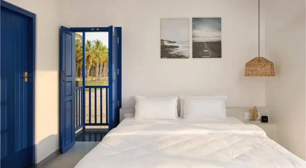 Minimalist white bedroom with crisp white bed linen, vibrant royal blue double doors opening to a balcony with coconut palm trees and beach view, framed coastal wall art, and a woven pendant light fixture.