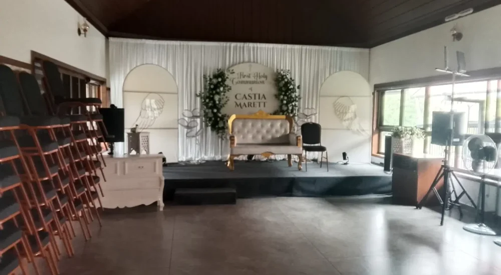 Arcadia Resort & Banquet ,First Holy Communion stage decoration with white backdrop and elegant seating inside Arcadia Resort banquet hall.