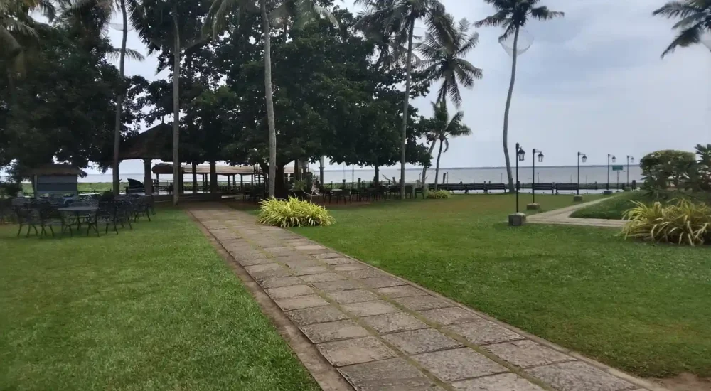 Intimate garden lawn and stone pathway near the traditional mandapam area with lush palm trees and backwater views at Backwater Ripples Hotel Kumarakom.
