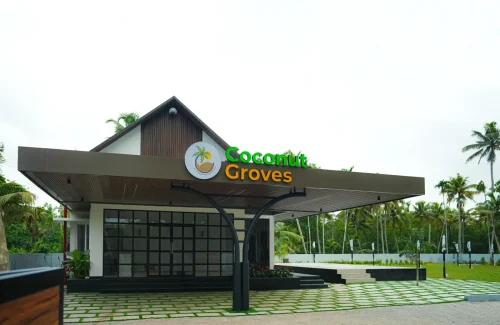 Exterior of Coconut Groves Convention Centre Kanjiramattom, showing the modern reception building with green and gold logo, a paved entrance, an outdoor stage, and a lawn surrounded by coconut palms.