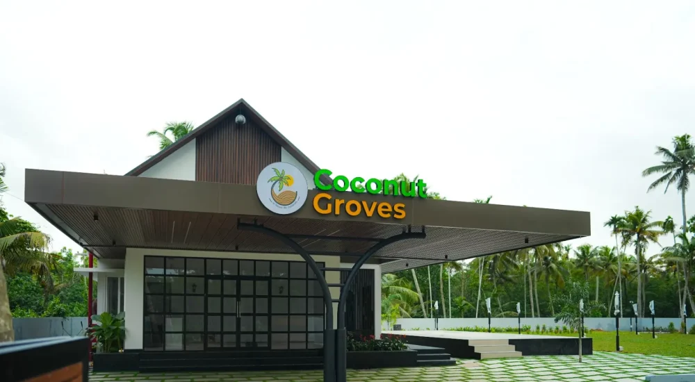Exterior of Coconut Groves Convention Centre Kanjiramattom, showing the modern reception building with green and gold logo, a paved entrance, an outdoor stage, and a lawn surrounded by coconut palms.