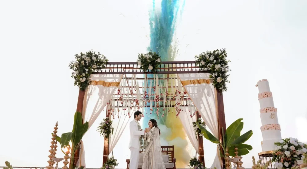 Bride and groom standing under outdoor wooden pergolas mandap with colorful blue smoke pyro effect at Rhythm Kumarakom backwaters