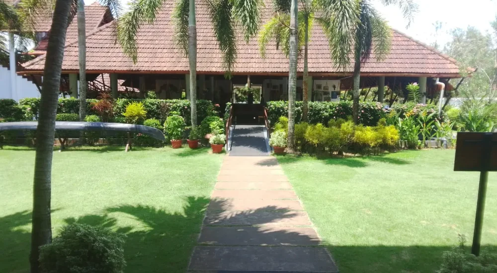 Stone walkway through green lawn leading to traditional wooden restaurant entrance at Rhythm Kumarakom resort