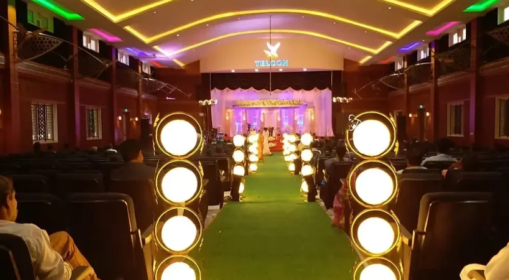 Modern indoor wedding reception decor by Melodia Events, showcasing a minimalist geometric backdrop with lush green foliage, white roses, structured pampas grass arrangements, and sleek LED linear lighting on a polished indoor stage.